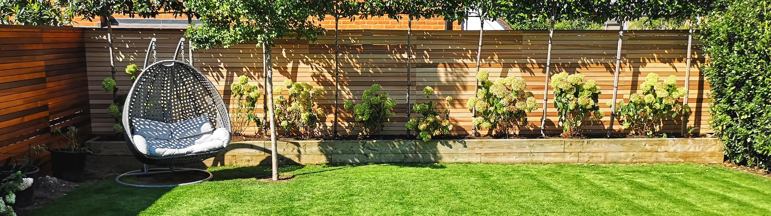 Backyard garden with green lawn, wooden fence, hydrangeas, and a hanging chair under a tree.