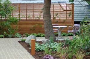 Garden patio with a round table, two chairs, a tree, and a slatted wooden fence.
