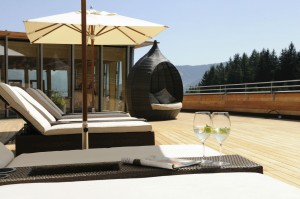 Sunlit rooftop terrace with loungers, umbrella, and two glasses on a table. Scenic view of trees and mountains.