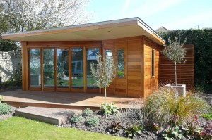 A modern wooden garden room with large glass doors, surrounded by plants and a small landscaped area.