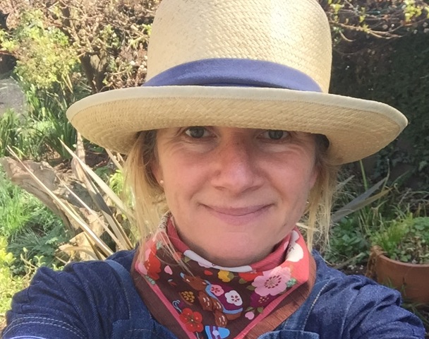 Woman in a straw hat and scarf, smiling outdoors in a garden setting.