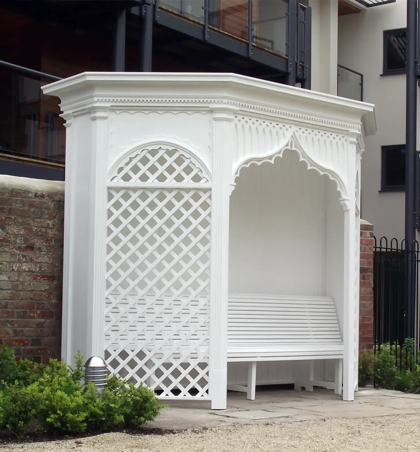 White decorative garden bench with lattice design under a roof, set against a brick wall.