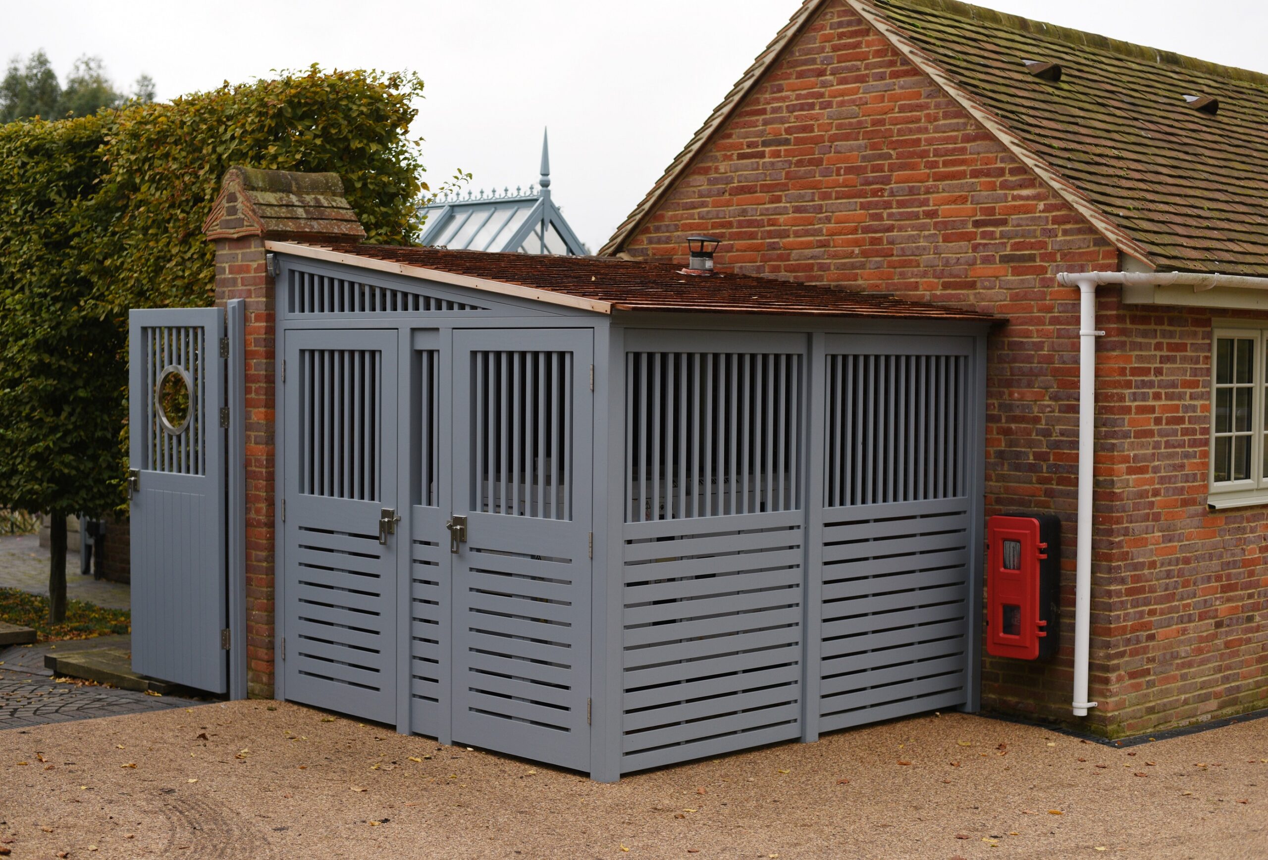 Gray wooden enclosure next to a brick building with a sloped roof and a red fire extinguisher on the wall.