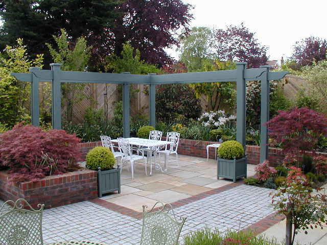 A garden patio with a metal pergola, white table, chairs, and various potted plants and shrubs.