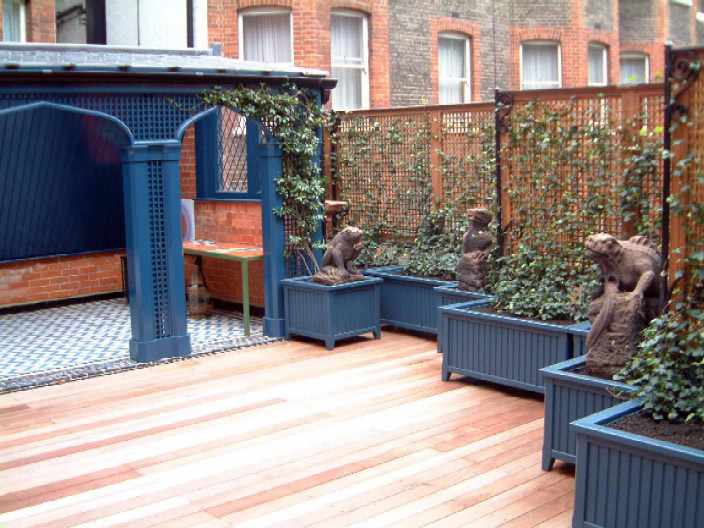 A patio with blue arched structure, wooden decking, and statues in planter boxes against a brick building backdrop.