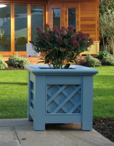 Blue lattice planter with plant, on patio, in front of a lawn and wooden garden building.