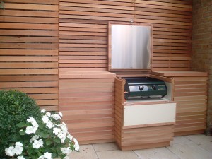 Outdoor wooden cabinet with a built-in gas grill, surrounded by plants and a wooden fence.