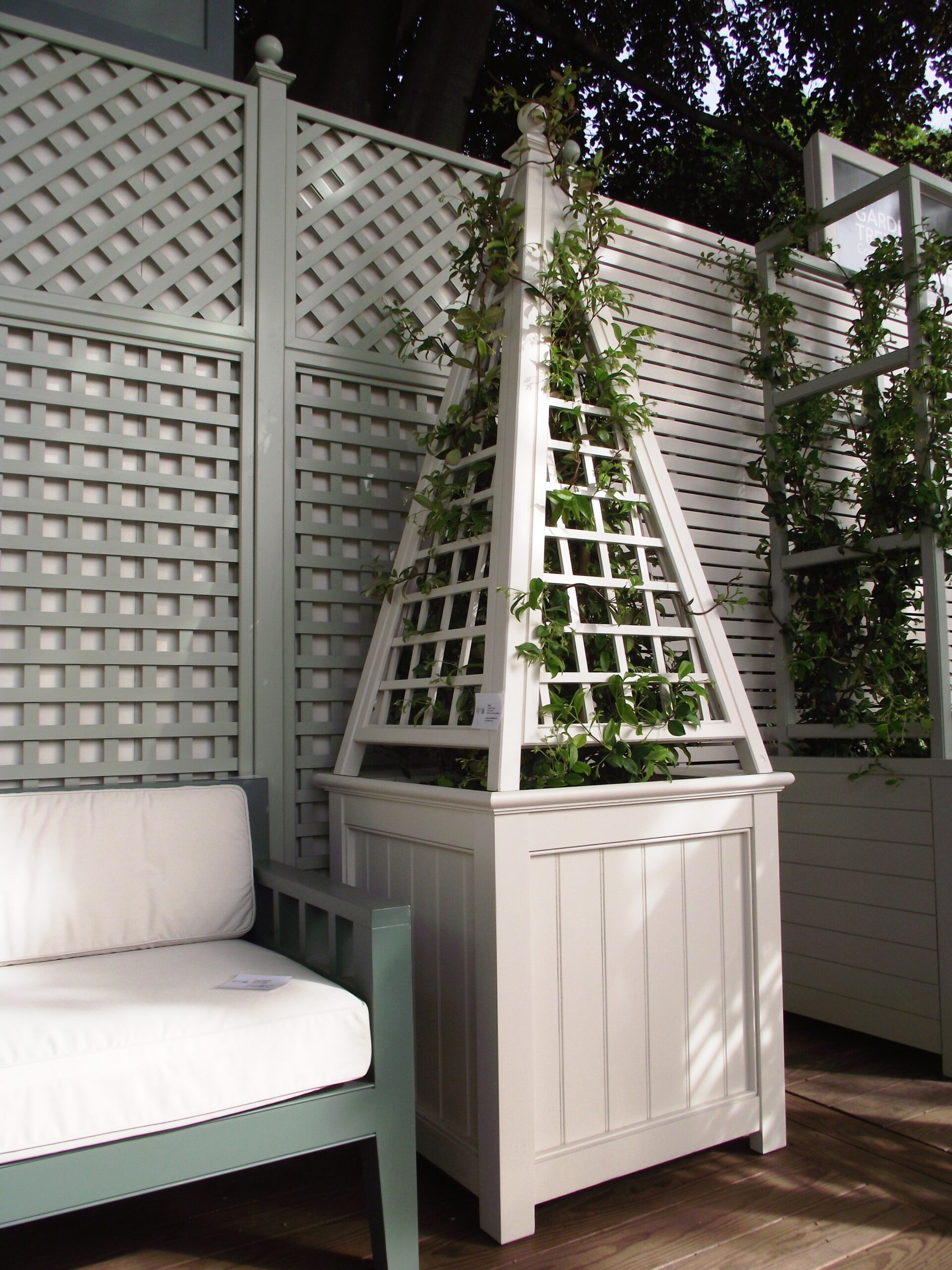 White trellis planter with climbing vine next to a white cushioned chair on a wooden deck.