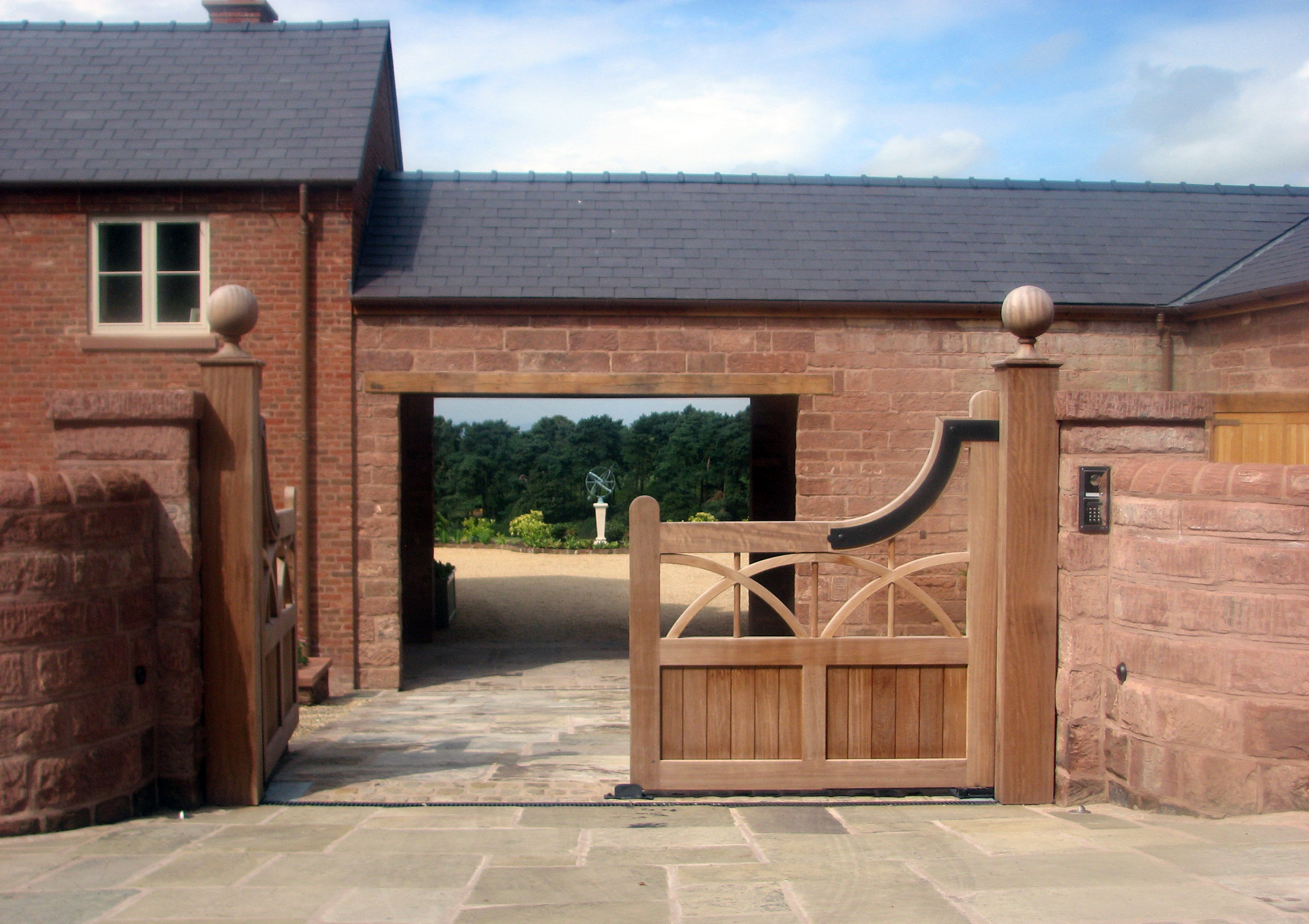 Wooden gate in a stone wall, leading to a courtyard with a brick building and tree-lined background.