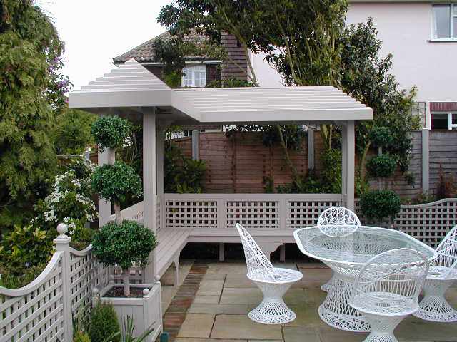 White outdoor pergola with lattice design, surrounded by greenery, featuring a white table and chairs on a stone patio.