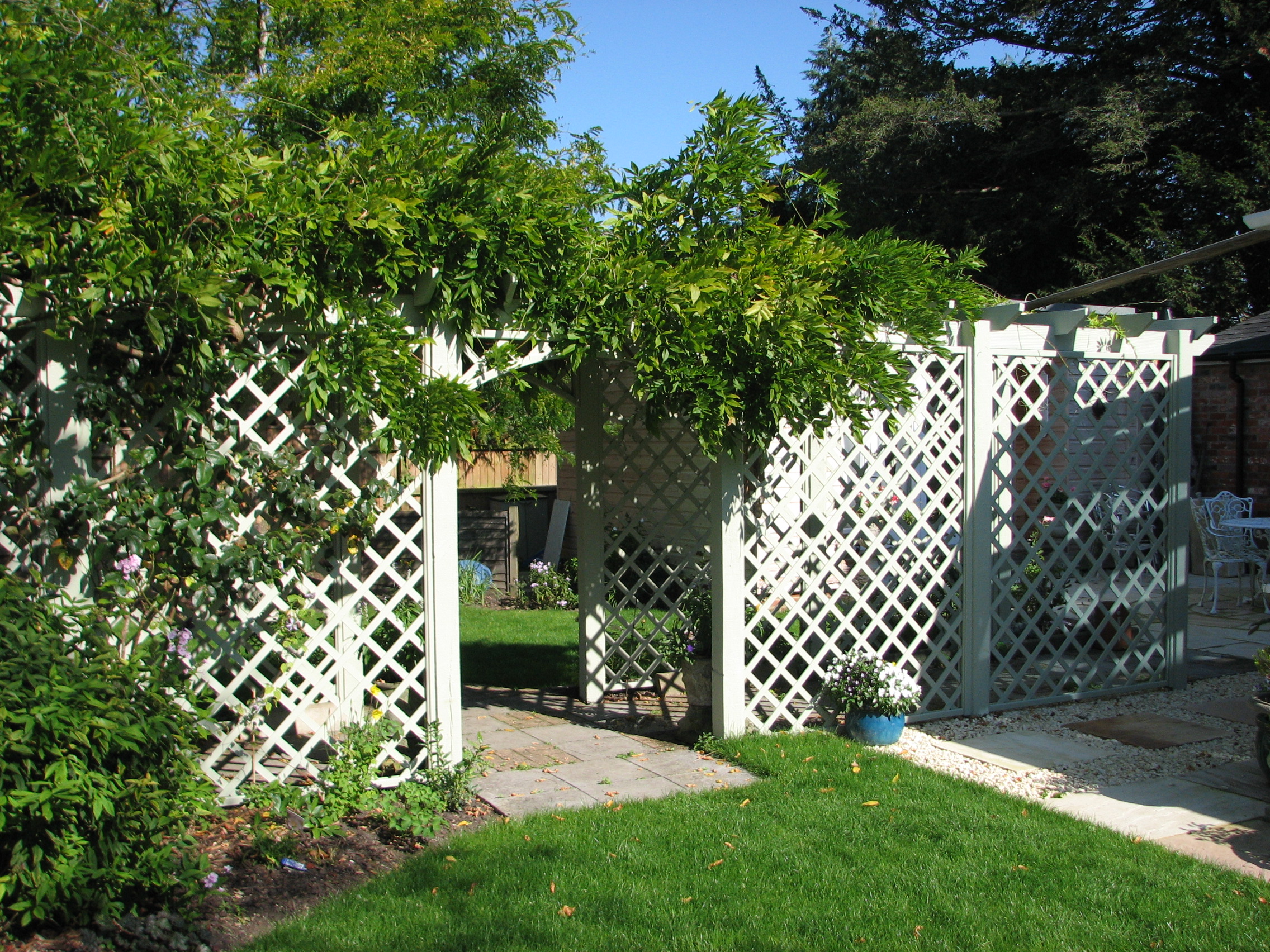 Garden with white lattice fence, green foliage, and a pathway leading to a sunlit outdoor area.