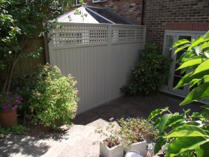 Small, paved backyard with plants and a white fenced corner next to a building with brick walls.