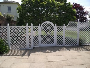 White lattice fence with an arched gate, set on a stone pathway, with trees and a house in the background.