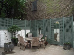 Patio with a round wooden table, chairs, potted plants, green lattice fence, and a brick wall backdrop.
