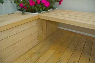 Corner of a wooden deck with integrated benches and pink flowering plants in a white planter.