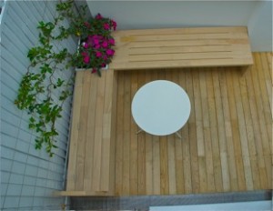 Wooden deck with a circular white table, L-shaped bench, and potted flowers in the corner.