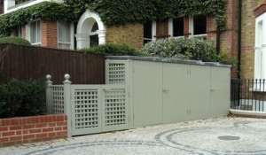 Green wooden storage shed with lattice accents outside a brick building with ivy.