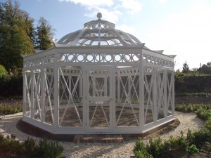 White octagonal garden structure with a domed roof, surrounded by gravel and plants, under a clear sky.