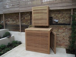 Wooden louvered enclosures cover outdoor units against a brick wall in a garden area.