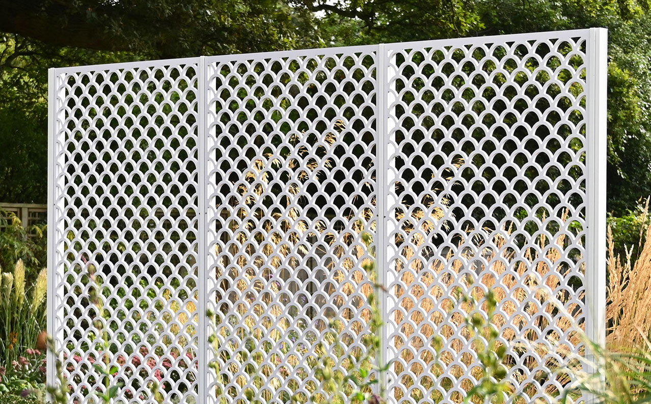 White decorative garden screen with a scallop pattern, set against a backdrop of greenery.