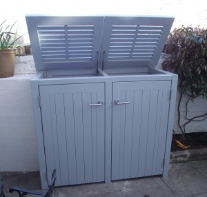 Gray outdoor storage cabinet with two compartments, each with a hinged lid and handle, placed on a stone patio.