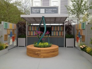 Garden display with plants, a wooden sculpture, colorful panels, and a sign overhead in an outdoor setting.