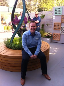 Man sitting on a circular wooden bench in a garden setting, with plants and latticework in the background.