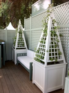 White trellises with climbing plants flank a bench against a lattice fence on a wooden deck.
