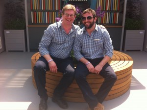 Two men sitting on a circular wooden bench, both wearing checked shirts, smiling at the camera.