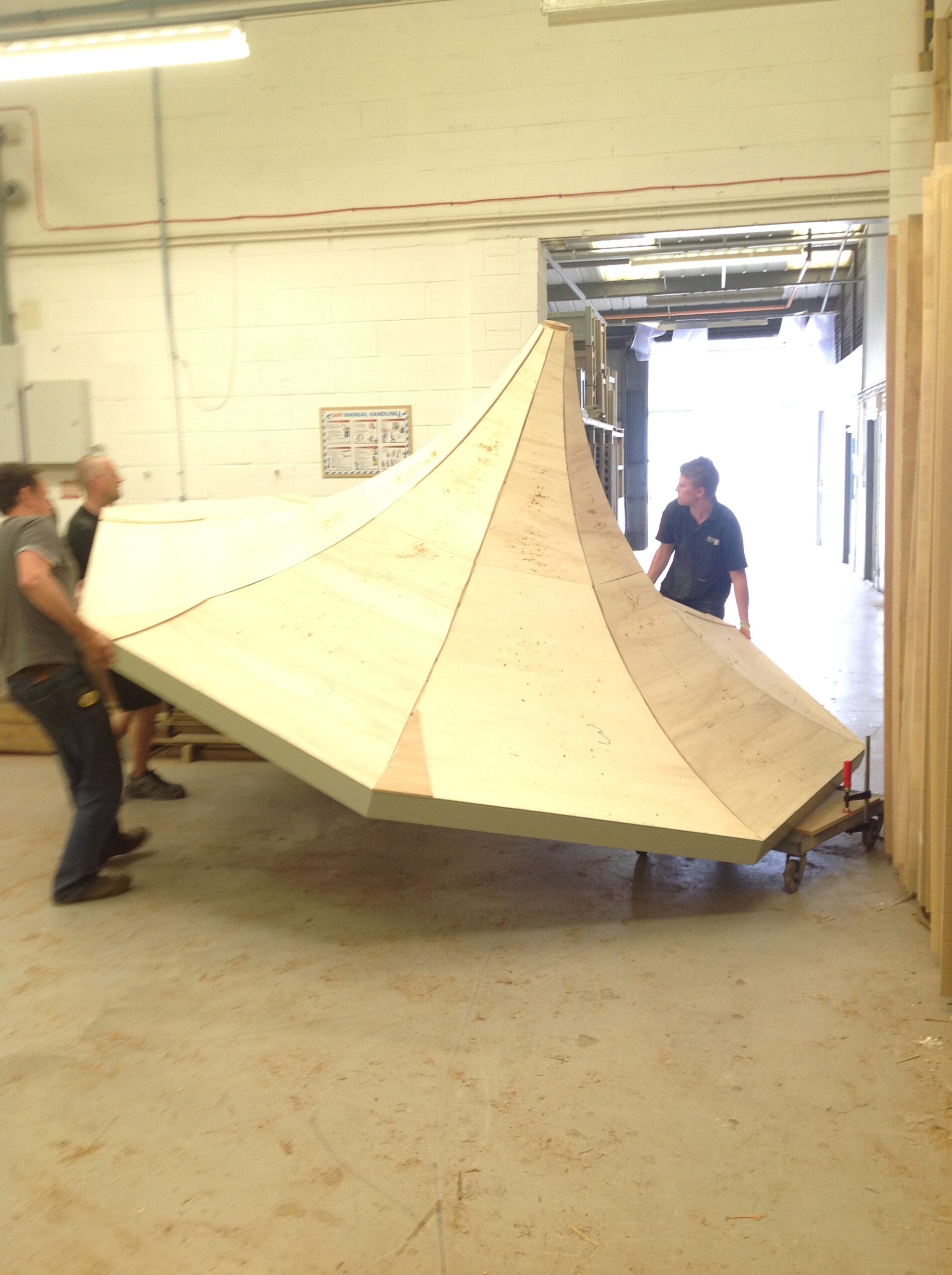 Three workers maneuver a large, geometrically shaped wooden structure through a doorway in a workshop.