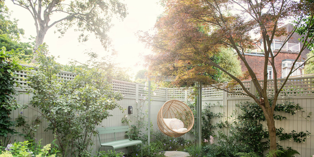 A tranquil garden with a hanging chair, surrounded by plants and trees, beside a wooden fence and a brick building.