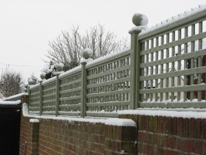 Snow-covered lattice fence on a brick wall, with bare trees in the background.
