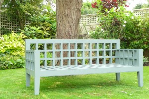 A gray wooden bench on a lawn, surrounded by trees and bushes, with a lattice fence in the background.