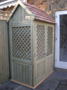 Wooden lattice storage shed with a sloped shingle roof, placed against a brick wall and next to a glass door.