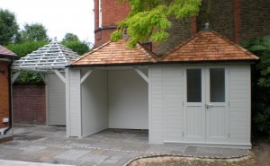 Wooden outbuilding with a shingled roof and attached pergola, situated on a stone patio near a brick residential building.