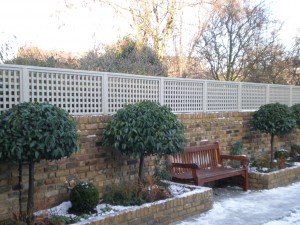 A wooden bench sits against a brick wall with trellis fencing, surrounded by trimmed shrubs in a snowy garden.