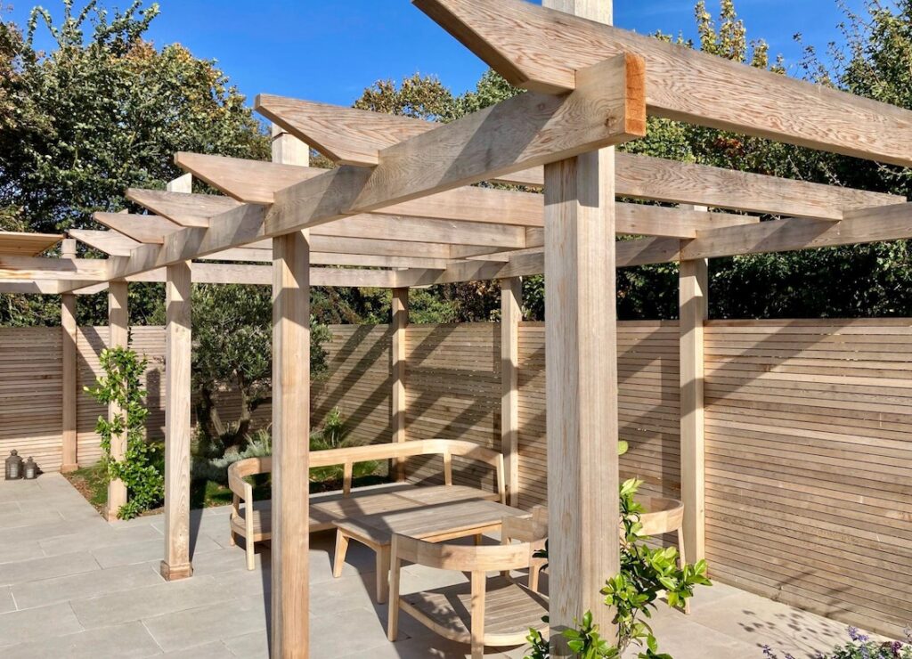 Wooden pergola with an L-shaped bench on a patio, surrounded by a wooden fence and trees.