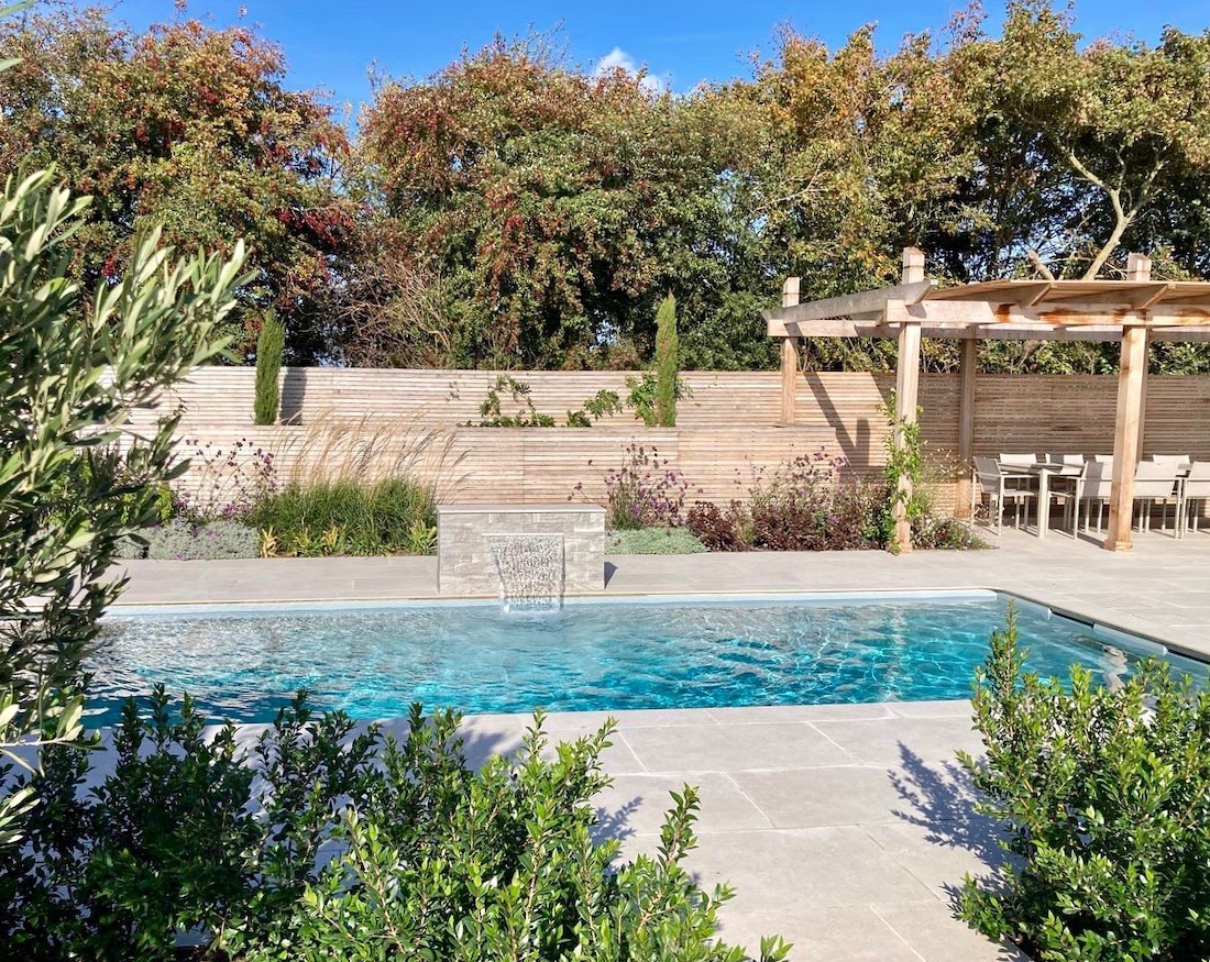 Outdoor pool with clear water, surrounded by greenery, a wooden pergola, and a dining area under a blue sky.