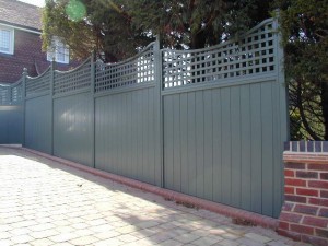 Tall green fence with lattice design on top, lining a paved pathway beside a brick building.