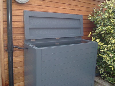 Gray outdoor storage box with an open lid, placed against a wooden wall near a pipe and leafy bush.