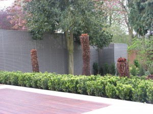 Trimmed trees and shrubs by a wooden deck, with a corrugated fence and taller trees in the background.
