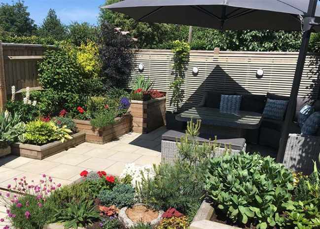 Patio with a sectional sofa, umbrella, and surrounding raised flower beds in a sunny garden.