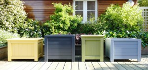 Four colorful square planters on a wooden deck with greenery and a wooden building in the background.