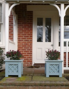 White door with glass panes, brick wall, porch, and two blue planters with flowers on stone steps.
