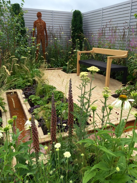 Garden with wooden bench, plants, flowers, small water feature, and human-shaped sculpture in a fenced area.