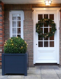 Decorated front door with a wreath, next to a lighted bush in a planter.