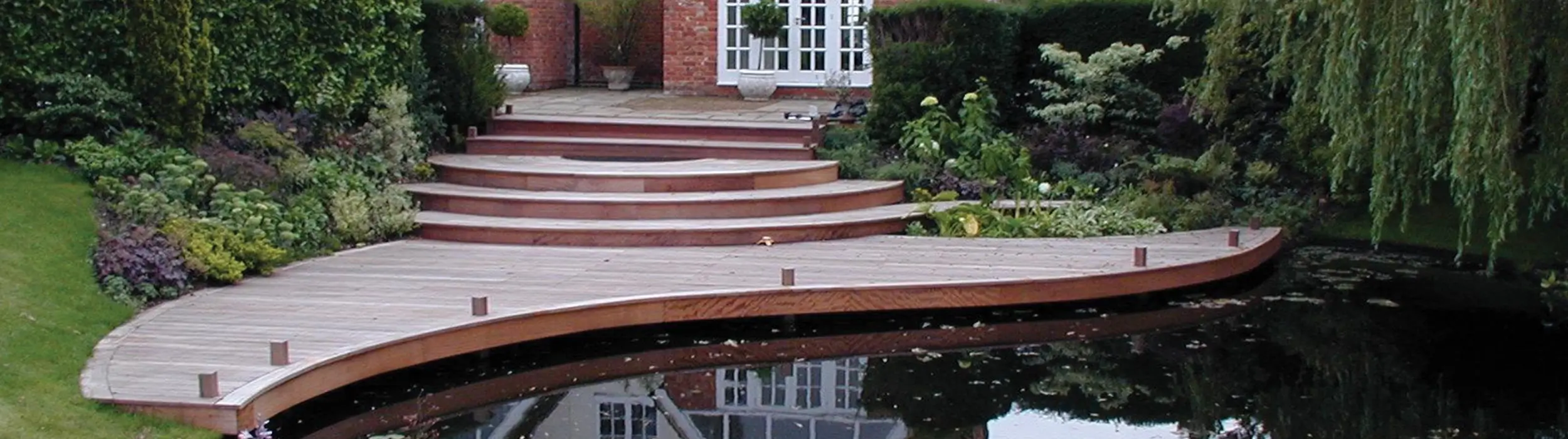 Curved wooden deck with steps, surrounded by greenery and a pond in a garden setting.