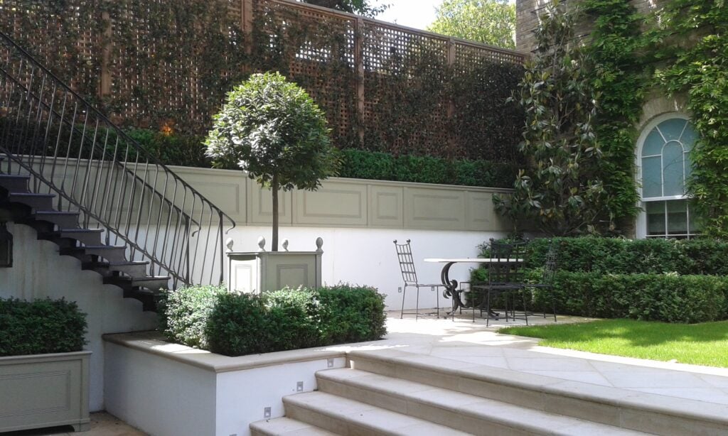 Garden patio with stairs, greenery, a table and chairs, and a small tree in a planter.