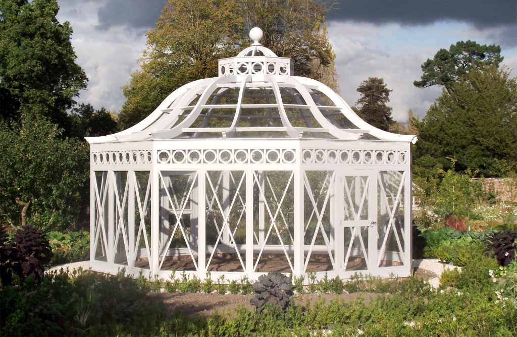 A white, ornate greenhouse with a domed roof in a garden setting under a cloudy sky.
