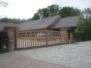 Wooden gate in front of a house with a stone driveway and trees in the background.